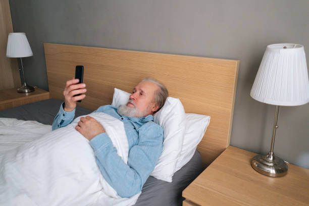 High-angle side view of handsome bearded senior adult male searching information about how to good health on internet, lying on bed in bedroom at home. Aged elderly man having video call at hospital.
